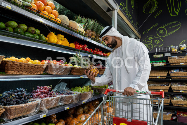 مجموعة متنوعة من الخضار الطازج ، رجل عربي خليجي إماراتي يرتدي الكندورة البيضاء يتفحص ثمرة ألانانس داخل أحد السوبرماركت، عربة تسوق تحتوي على منتجات غذائية ومنزلية، الاستفادة من الخصم علي المنتجات