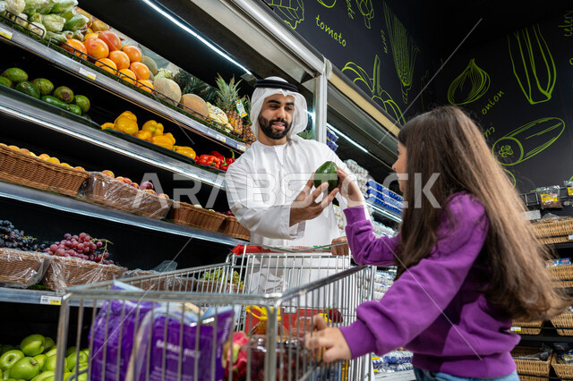 Buying essential household needs, helping and teaching children to buy from the market, advising the daughter in buying household goods and supplies, Emirati national agricultural crops, an Emirati Gulf Arab father wearing a white kandura giving his daughter a green pear