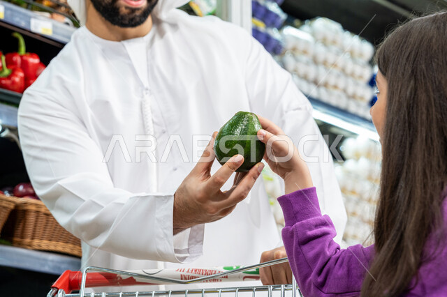مساعدة وتعليم الأبناء الشراء من الماركت، ترتيب المشتريات داخل عربة التسوق، إستشارة الإبنة في شراء السلع والمستلزمات المنزلي، محاصيل زراعية وطنية اماراتية، صورة مقربه  لأب عربي خليجي اماراتي يرتدي الكندورة البيضاء يقوم بإعطاء ابنته ثمرة كمثره خضراء