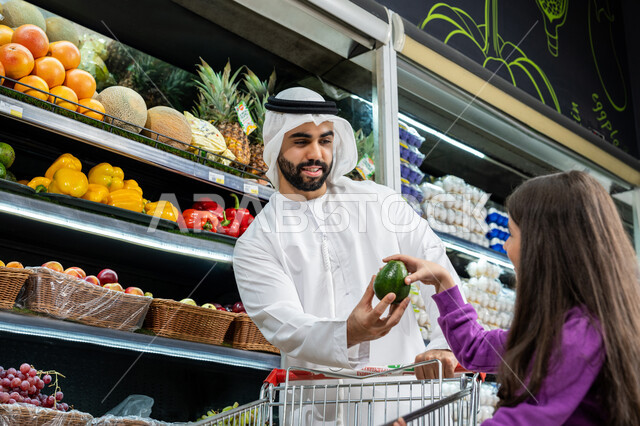 قضاء أوقات ممتعه مع الأبناء، ترتيب المشتريات داخل عربة التسوق، إستشارة الإبنة في شراء السلع والمستلزمات المنزلي، محاصيل زراعية وطنية اماراتية، اب عربي خليجي اماراتي يرتدي الكندورة البيضاء يقوم بإعطاء ابنته ثمرة كمثره خضراء