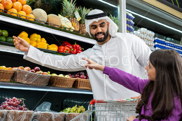 Browsing through different items, arranging purchases in the shopping cart, consulting the daughter in purchasing goods and household supplies, Emirati national agricultural crops, an Emirati Gulf Arab father wearing a white kandura choosing colorful peppers in the supermarket