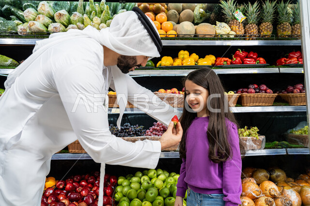 محاصيل زراعية وطنية اماراتية، تعليم الأبناء أنواع الفاكهه والخضروات المعروضه على أرفف الثلاجه، إختيار أفضل الفواكه الطازجة، إستشارة الإبنة في شراء السلع والمستلزمات المنزلية، اب عربي خليجي اماراتي يرتدي الكندورة البيضاء يقوم بإعطاء ابنته ثمره فراوله  في السوبرماركت