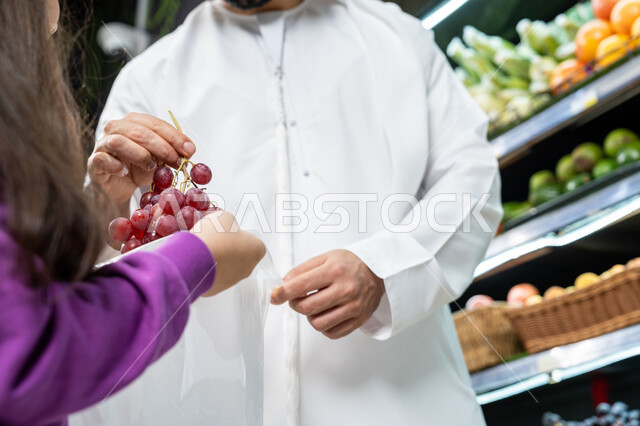 إختيار أفضل الفواكه الطازجة، إستشارة الإبنة في شراء السلع والمستلزمات المنزلية، صورة مقربه لأب عربي خليجي اماراتي يرتدي الكندورة البيضاء يقوم بإعطاء ابنته ثمره عنب طازجه في السوبرماركت، تعليم الأبناء أنواع الفاكهه والخضروات المعروضه على أرفف الثلاجه في الماركت