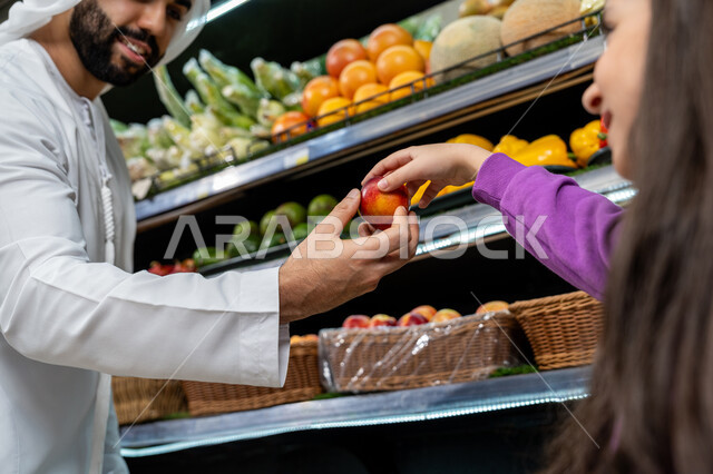 Having different purchasing and consumption options in the store, the joy of shopping with the children in the supermarket, a close-up photo of an Emirati Gulf Arab man giving his daughter a fresh red apple with gestures of happiness and pleasure, local Emirati national agricultural products and crops