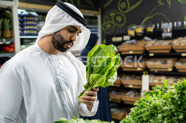 متعة التسوق وشراء السلع المختلفة، الوعي الغذائي والاهتمام بالتفاصيل أثناء التسوق، التبضع واختيار الخضروات الورقية الطازجة داخل السوبرماركت،صورة مقربه لرجل عربي خليجي إماراتي يرتدي الكندورة البيضاء يتفحص باقة من الخس للتأكد من جودتها
