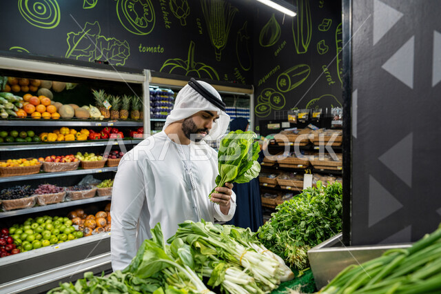 الوعي الغذائي والاهتمام بالتفاصيل أثناء التسوق، الإستفادة من العرض و التخفيض على السلع الغذائية، التبضع واختيار الخضروات الورقية الطازجة داخل السوبرماركت، رجل عربي خليجي إماراتي يرتدي الكندورة البيضاء يتفحص باقة من الخس للتأكد من جودتها