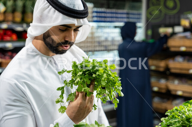 التبضع واختيار الخضروات الورقية الطازجة داخل السوبرماركت، صورة مقربة لرجل عربي خليجي إماراتي يرتدي الكندورة البيضاء يتفحص باقة من البقدونس للتأكد من جودتها، امرأة خليجية تتجول عند الرفوف، الوعي الغذائي والاهتمام بالتفاصيل أثناء التسوق