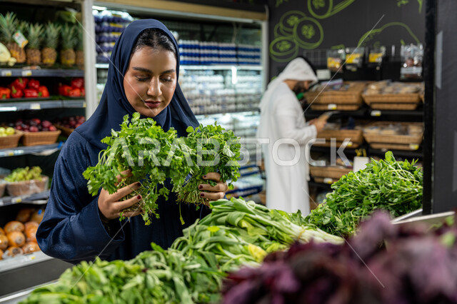 Paying attention to the quality of food products while shopping at the supermarket, the pleasure of shopping and choosing fresh leafy vegetables, an Emirati Gulf Arab couple wandering inside the vegetable section, a woman carefully selecting fresh parsley, cooperation and distribution of roles within the Gulf family
