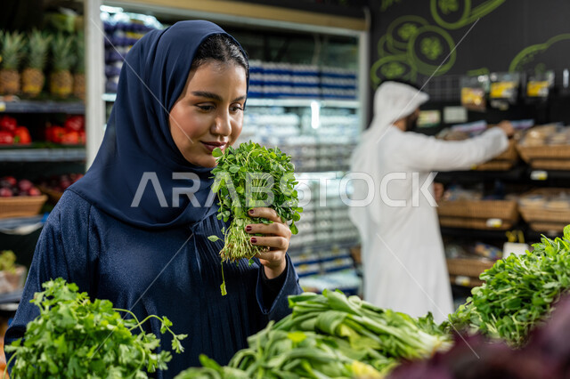 متعة التبضع واختيار الخضروات الورقية الطازجة، الاهتمام بجودة المنتجات الغذائية أثناء التسوق في السوبرماركت، زوجان عربيان خليجيان إماراتيان يتجولان داخل قسم الخضروات، امراءة تقوم باختيار البقدونس الطازج بعناية، التعاون وتوزيع الأدوار داخل الأسرة الخليجية