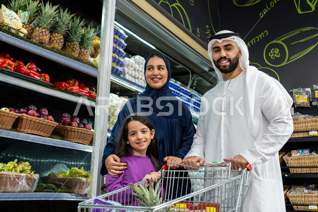 Enjoying discounts, offers and reductions on products, cooperating in purchasing various goods and food items in the supermarket, an Arab Gulf Emirati couple spending enjoyable times shopping and choosing household supplies with their daughter, helping and teaching the children how to shop