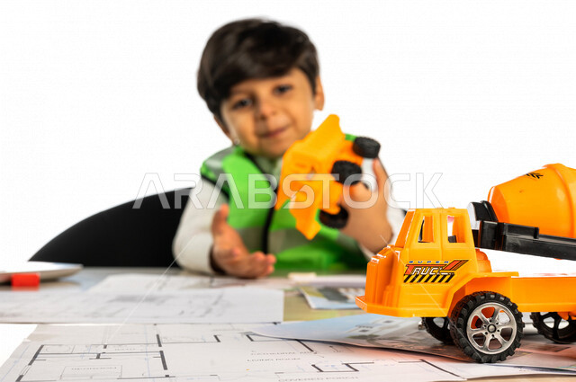 بورتريه لفتى عربي خليجي سعودي داخل مكتب هندسي، بيده لعبة شاحنة خلاطة أسمنت صغيرة، إستخدام جهاز الحاسوب المحمول، أدوات ومعدات هندسية، يرتدي سترة الحماية الخاصة بالعمل ، مخططات وتصماميم هندسية، مهنة مستقبلية، خلفية بيضاء، مفهوم الهندسة المعمارية
