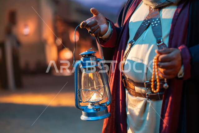 Heritage of the Kingdom of Saudi Arabia, a traditional folk masterpiece, the anniversary of the founding of the first Saudi state, February 22, the day of our beginnings, 1727 AD, an old antique metal lantern, an old-fashioned lamp made of copper, a close-up of a Saudi Arabian Gulf man wearing a belt holding a prayer bead and a traditional lantern at night