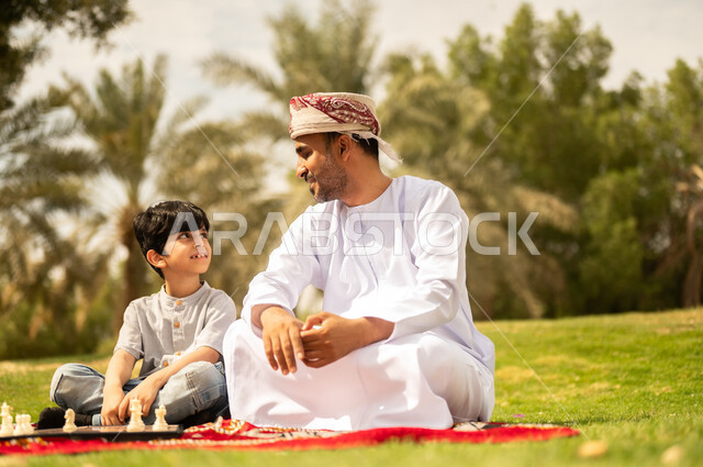 نزهه في الهواء الطلق، رجل عماني يرتدي العمامة والدشداشة يلعب مع ابنه لعبة الضامة، الجلوس في الطبيعة الخضراء، مفهوم التحدي والتركيز، عائلة عربية خليجية عمانية تقضي أوقات ممتعة في إحدى الحدائق في سلطنة عمان، أنشطة عائلية في الأجواء الصيفية