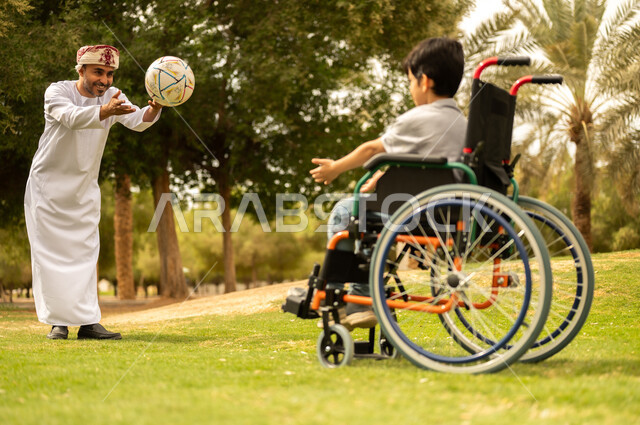 Playing football with gestures of joy and happiness, practicing recreational activities and spending fun times with children, providing supplies for the disabled, an Arab Gulf Omani father wearing a turban and dishdasha playing with a boy sitting in a wheelchair in the park, nature background