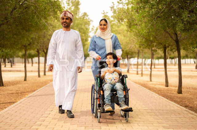الاستعداد للعب بكرة القدم، ممرضة عربية خليجية عمانية محجبة تقوم بمساعدة ولد يجلس على الكرسي المتحرك، نمط الحياة في سلطنة عمان، رعاية ذوي الاحتياجات الخاصة، تأمين ادوات تساعد أصحاب الهمم على التنقل، أب يستمتع بالتنزه في إحدى الحدائق في الأجواء الصيفية مع ابنه