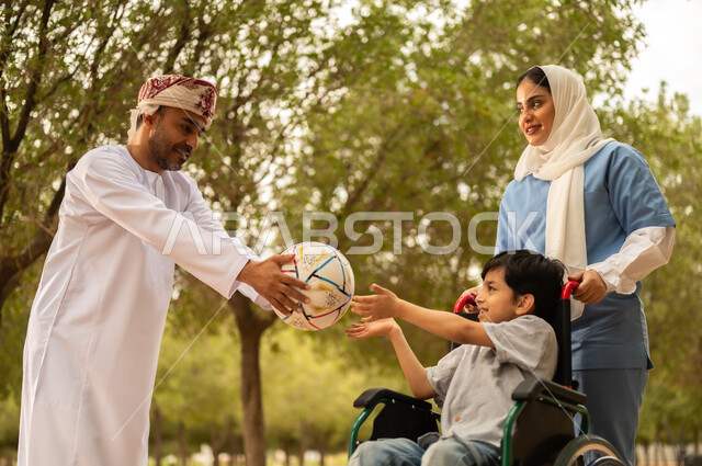 رعاية ذوي الاحتياجات الخاصة، تأمين ادوات تساعد أصحاب الهمم على التنقل، أب يستمتع بالتنزه في إحدى الحدائق في الأجواء الصيفية مع ابنه، الاستعداد للعب بكرة القدم، ممرضة عربية خليجية عمانية محجبة تقوم بمساعدة ولد يجلس على الكرسي المتحرك، نمط الحياة في سلطنة عمان