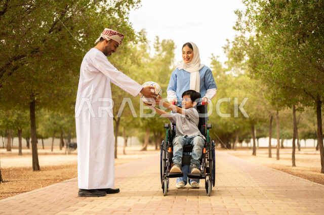 رعاية ذوي الاحتياجات الخاصة، تأمين ادوات تساعد أصحاب الهمم على التنقل، أب يستمتع بالتنزه في إحدى الحدائق في الأجواء الصيفية مع ابنه، الاستعداد للعب بكرة القدم، ممرضة عربية خليجية عمانية محجبة تقوم بمساعدة ولد يجلس على الكرسي المتحرك، نمط الحياة في سلطنة عمان