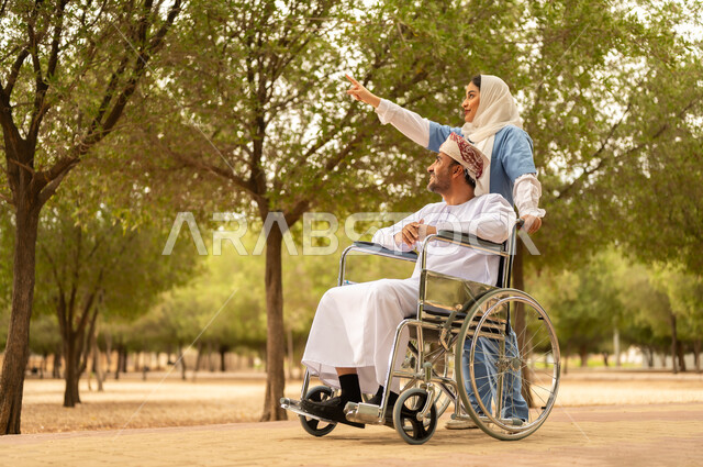 تأمين ادوات تساعد أصحاب الهمم على التنقل والحركة، الاستمتاع  بالأجواء الصيفيه المشمسة، التنزه في احدى الحدائق، ممرضة عربية خليجية عمانية محجبة تقوم بمساعدة رجل يرتدي الدشداشة والعمامة يجلس على الكرسي المتحرك، الرعاية والاهتمام بذوي الاحتياجات