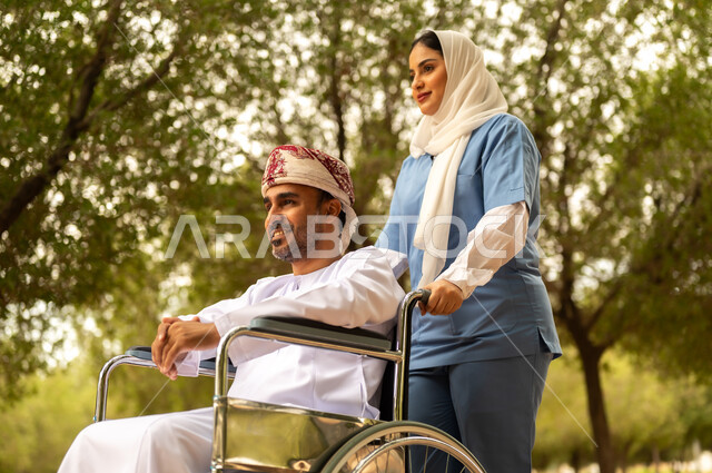 Caring for and paying attention to people with special needs, providing tools to help people with disabilities move around, enjoying the sunny summer weather, taking a walk in a park, a veiled Gulf Arab Omani nurse helping a man wearing a dishdasha and turban sitting in a wheelchair
