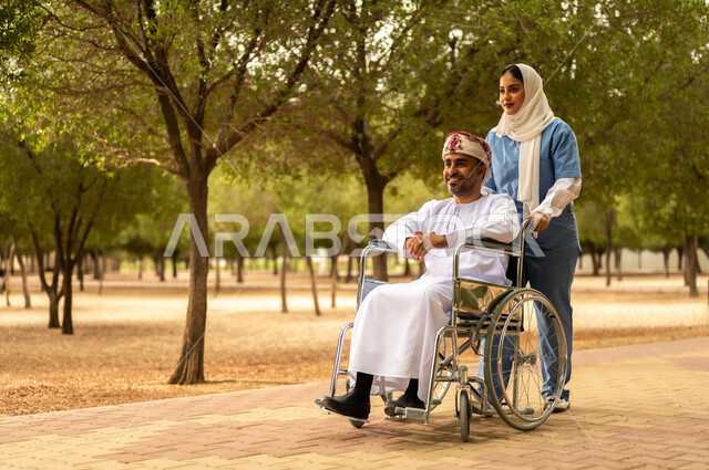 الرعاية والاهتمام بذوي الاحتياجات، تأمين ادوات تساعد أصحاب الهمم على التنقل والحركة، الاستمتاع  بالأجواء الصيفيه المشمسة، التنزه في احدى الحدائق، ممرضة عربية خليجية عمانية محجبة تقوم بمساعدة رجل يرتدي الدشداشة والعمامة يجلس على الكرسي المتحرك