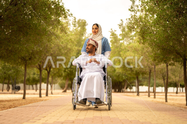 تأمين ادوات تساعد أصحاب الهمم على التنقل والحركة، الاستمتاع  بالأجواء الصيفيه المشمسة، التنزه في احدى الحدائق، ممرضة عربية خليجية عمانية محجبة تقوم بمساعدة رجل يرتدي الدشداشة والعمامة يجلس على الكرسي المتحرك، الرعاية والاهتمام بذوي الاحتياجات