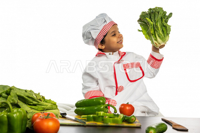 Portrait of a Saudi Arabian Gulf boy, wearing a special chef uniform, standing in front of a table with a group of fresh vegetables, holding in his hand fresh lettuce, a young chef, a future profession, preparing salads and healthy dishes, white backgroun