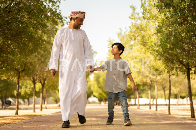 أجواء أسرية ممتعة، قضاء أوقات الإجازة مع الابناء، الترابط الأسري و الاستمتاع بالعطلة مع العائلة، مفهوم الرابطة الأبوية، ايماءات السعادة و السرور، أب عربي خليجي عماني يرتدي الدشداشة والعمامة يلعب ويتنزه في الهواء الطلق برفقة ولده، خلفية الطبيعة
