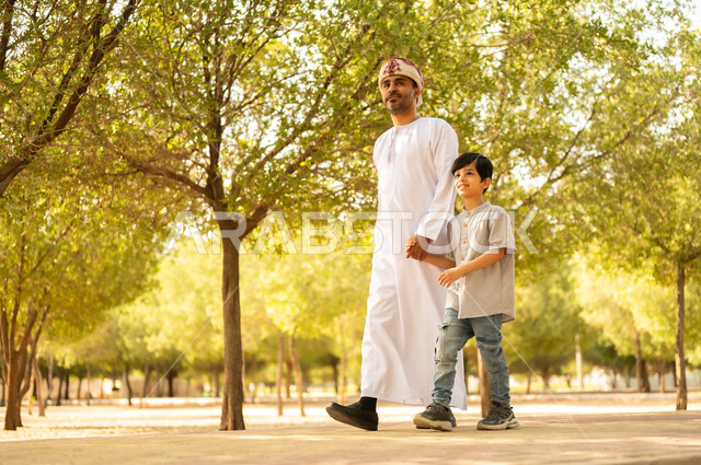 أجواء أسرية ممتعة، قضاء أوقات الإجازة مع الابناء، الترابط الأسري و الاستمتاع بالعطلة مع العائلة، مفهوم الرابطة الأبوية، ايماءات السعادة و السرور، أب عربي خليجي عماني يرتدي الدشداشة والعمامة يلعب ويتنزه في الهواء الطلق برفقة ولده، خلفية الطبيعة
