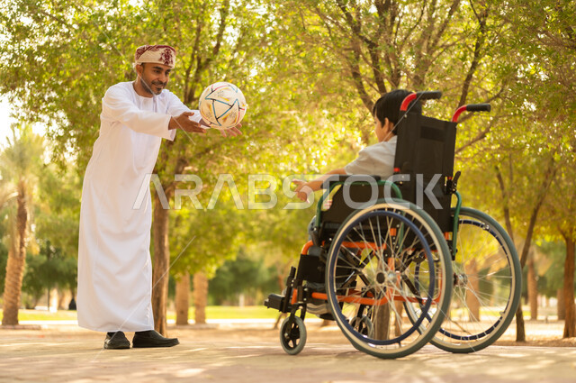 اللعب بكرة القدم بإيماءات الفرح والسرور، ممارسة الأنشطة الترفيهية وقضاء أوقات متعة مع الأبناء, توفير المستلزمات الخاصة بالمعاقين، أب عربي خليجي عماني يرتدي العمامة والدشداشة يلعب مع ولد يجلس على كرسي متحرك في الحديقة, خلفية الطبيعة