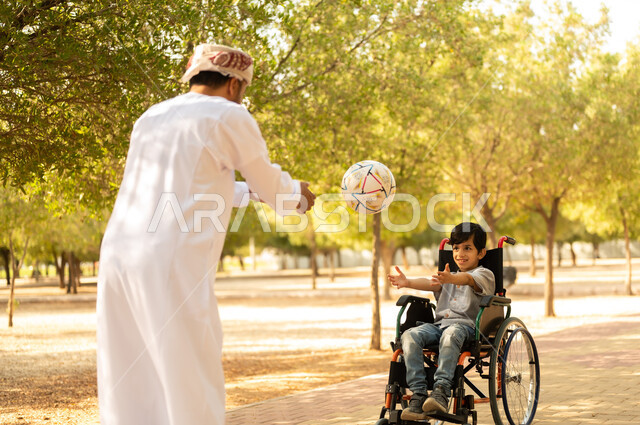 اللعب بكرة القدم بإيماءات الفرح والسرور، ممارسة الأنشطة الترفيهية وقضاء أوقات متعة مع الأبناء, توفير المستلزمات الخاصة بالمعاقين، أب عربي خليجي عماني يرتدي العمامة والدشداشة يلعب مع ولد يجلس على كرسي متحرك في الحديقة, خلفية الطبيعة