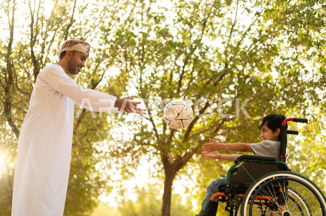 Playing football with gestures of joy and happiness, practicing recreational activities and spending fun times with children, providing supplies for the disabled, an Arab Gulf Omani father wearing a turban and dishdasha playing with a boy sitting in a wheelchair in the park, nature background