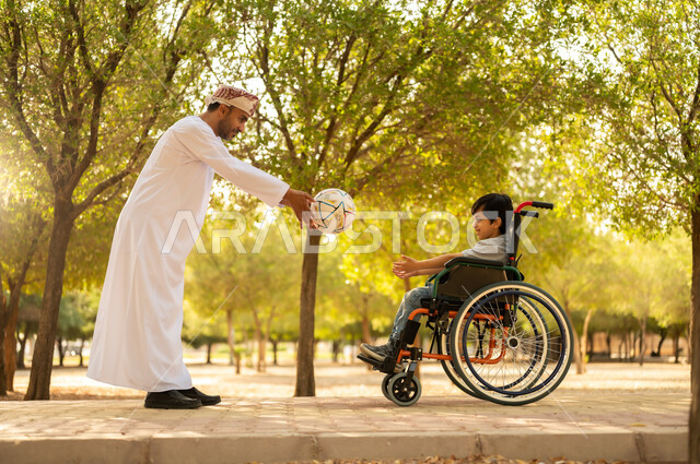 اللعب بكرة القدم بإيماءات الفرح والسرور، ممارسة الأنشطة الترفيهية وقضاء أوقات متعة مع الأبناء, توفير المستلزمات الخاصة بالمعاقين، أب عربي خليجي عماني يرتدي العمامة والدشداشة يلعب مع ولد يجلس على كرسي متحرك في الحديقة, خلفية الطبيعة