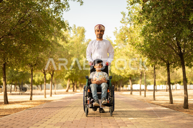 Providing for the needs of people of determination in the Sultanate of Oman, community interest and support for people with special needs, an Arab Gulf Omani father wearing a turban and a dishdasha walking with his disabled son, sitting in a wheelchair and spending time in the park with the family, a nature background