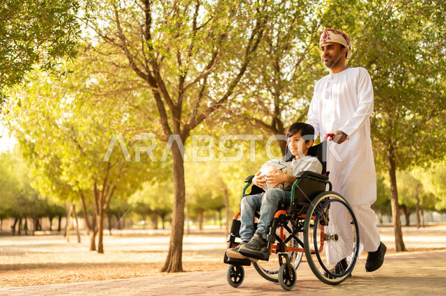 تأمين احتياجات أصحاب الهمم في سلطنة عمان، اهتمام ودعم المجتمع لذوي الاحتياجات الخاصه، أب عربي خليجي عماني يرتدي العمامة والدشداشة يتنزه مع ابنه المقعد، الجلوس على كرسي متحرك وقضاء الوقت في المنتزه مع العائلة، خلفية الطبيعة