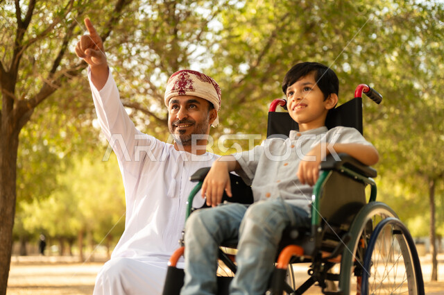 اهتمام ودعم المجتمع لذوي الاحتياجات الخاصه، تأمين احتياجات أصحاب الهمم في سلطنة عمان، أب عربي خليجي عماني يرتدي العمامة والدشداشة يتنزه مع ابنه المقعد، الجلوس على كرسي متحرك وقضاء الوقت في المنتزه مع العائلة، خلفية الطبيعة