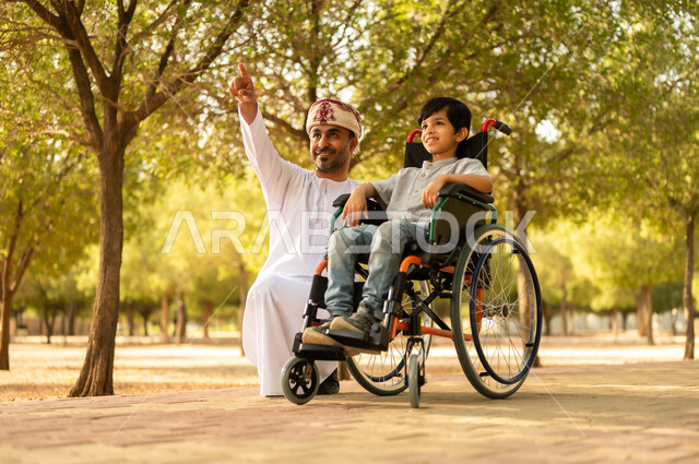 تأمين احتياجات أصحاب الهمم في سلطنة عمان، اهتمام ودعم المجتمع لذوي الاحتياجات الخاصه، أب عربي خليجي عماني يرتدي العمامة والدشداشة يتنزه مع ابنه المقعد، الجلوس على كرسي متحرك وقضاء الوقت في المنتزه مع العائلة، خلفية الطبيعة