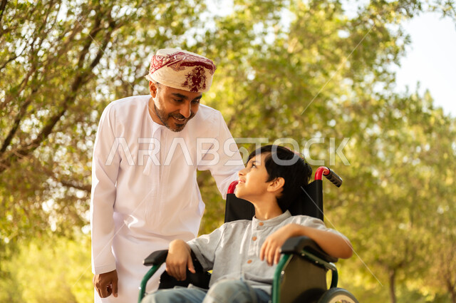 تأمين احتياجات أصحاب الهمم في سلطنة عمان، اهتمام ودعم المجتمع لذوي الاحتياجات الخاصه، أب عربي خليجي عماني يرتدي العمامة والدشداشة يتنزه مع ابنه المقعد، الجلوس على كرسي متحرك وقضاء الوقت في المنتزه مع العائلة، خلفية الطبيعة