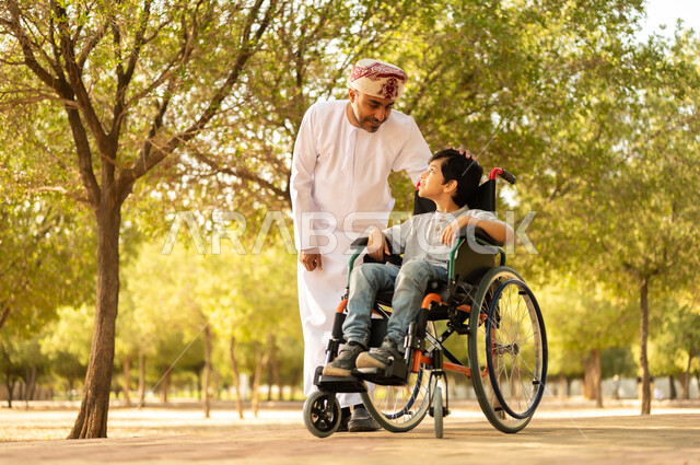 اهتمام ودعم المجتمع لذوي الاحتياجات الخاصه، تأمين احتياجات أصحاب الهمم في سلطنة عمان، أب عربي خليجي عماني يرتدي العمامة والدشداشة يتنزه مع ابنه المقعد، الجلوس على كرسي متحرك وقضاء الوقت في المنتزه مع العائلة، خلفية الطبيعة