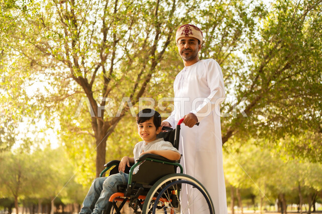 Providing for the needs of people of determination in the Sultanate of Oman, community interest and support for people with special needs, an Arab Gulf Omani father wearing a turban and a dishdasha walking with his disabled son, sitting in a wheelchair and spending time in the park with the family, a nature background