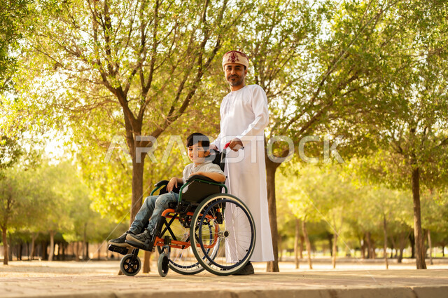 تأمين احتياجات أصحاب الهمم في سلطنة عمان، اهتمام ودعم المجتمع لذوي الاحتياجات الخاصه، أب عربي خليجي عماني يرتدي العمامة والدشداشة يتنزه مع ابنه المقعد، الجلوس على كرسي متحرك وقضاء الوقت في المنتزه مع العائلة، خلفية الطبيعة