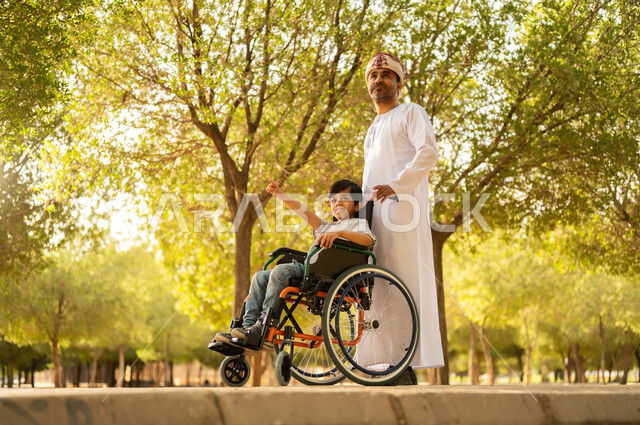 اهتمام ودعم المجتمع لذوي الاحتياجات الخاصه، تأمين احتياجات أصحاب الهمم في سلطنة عمان، أب عربي خليجي عماني يرتدي العمامة والدشداشة يتنزه مع ابنه المقعد، الجلوس على كرسي متحرك وقضاء الوقت في المنتزه مع العائلة، خلفية الطبيعة