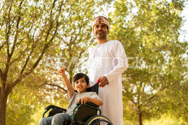 تأمين احتياجات أصحاب الهمم في سلطنة عمان، اهتمام ودعم المجتمع لذوي الاحتياجات الخاصه، أب عربي خليجي عماني يرتدي العمامة والدشداشة يتنزه مع ابنه المقعد، الجلوس على كرسي متحرك وقضاء الوقت في المنتزه مع العائلة، خلفية الطبيعة