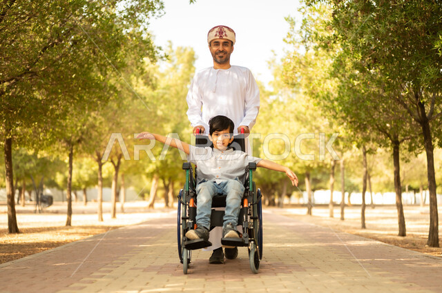 تأمين احتياجات أصحاب الهمم في سلطنة عمان، اهتمام ودعم المجتمع لذوي الاحتياجات الخاصه، أب عربي خليجي عماني يرتدي العمامة والدشداشة يتنزه مع ابنه المقعد، الجلوس على كرسي متحرك وقضاء الوقت في المنتزه مع العائلة، خلفية الطبيعة