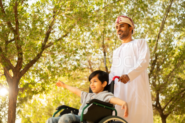 اهتمام ودعم المجتمع لذوي الاحتياجات الخاصه، تأمين احتياجات أصحاب الهمم في سلطنة عمان، أب عربي خليجي عماني يرتدي العمامة والدشداشة يتنزه مع ابنه المقعد، الجلوس على كرسي متحرك وقضاء الوقت في المنتزه مع العائلة، خلفية الطبيعة