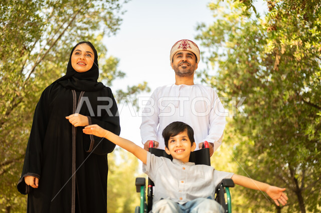 ولد عماني مقعد يجلس على كرسي متحرك في أحد المنتزهات، منح مرضى ذوي الإعاقة الحركية طاقة ايجابية، شعور ذوي الهمم بالراحه والطمأنينه في بيئتهم، عائلة عربيه خليجيه عمانية برفقه ابنها في إحدى الحدائق للاستمتاع بالهواء الطلق
