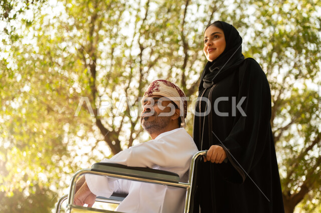 الاستمتاع وقضاء وقت ممتع بالهواء الطلق, العطف والحب بين أفراد الأسرة, امرأة عربية خليجية عمانية محجبة ترتدي العباءة تتنزه في الحديقة برفقة زوجها المقعد, شعور ذوي الهمم بالراحة والطمأنينة في بيئتهم, مساعدة مرضى الإعاقة الحركية على التنقل بسهولة