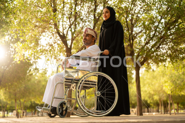 العطف والحب بين أفراد الأسرة, امرأة عربية خليجية عمانية محجبة ترتدي العباءة تتنزه في الحديقة برفقة زوجها المقعد, شعور ذوي الهمم بالراحة والطمأنينة في بيئتهم, مساعدة مرضى الإعاقة الحركية على التنقل بسهولة، الاستمتاع وقضاء وقت ممتع بالهواء الطلق