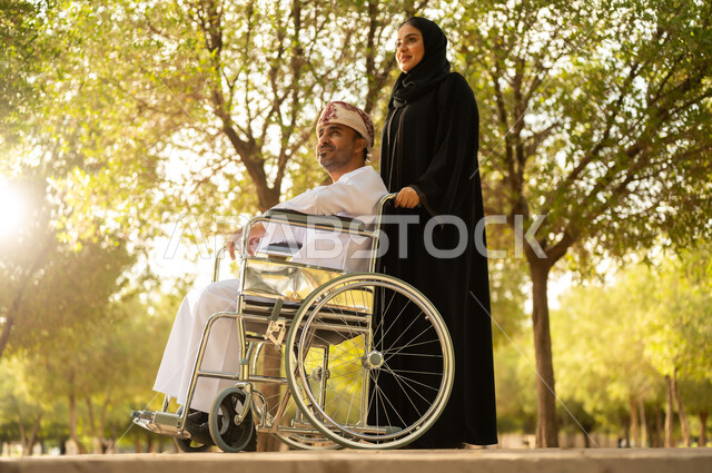 الاستمتاع وقضاء وقت ممتع بالهواء الطلق, العطف والحب بين أفراد الأسرة, امرأة عربية خليجية عمانية محجبة ترتدي العباءة تتنزه في الحديقة برفقة زوجها المقعد, شعور ذوي الهمم بالراحة والطمأنينة في بيئتهم, مساعدة مرضى الإعاقة الحركية على التنقل بسهولة