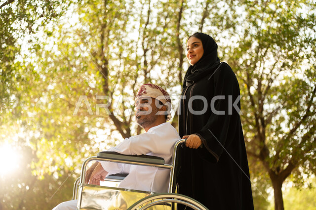 الاستمتاع وقضاء وقت ممتع بالهواء الطلق, العطف والحب بين أفراد الأسرة, امرأة عربية خليجية عمانية محجبة ترتدي العباءة تتنزه في الحديقة برفقة زوجها المقعد, شعور ذوي الهمم بالراحة والطمأنينة في بيئتهم, مساعدة مرضى الإعاقة الحركية على التنقل بسهولة