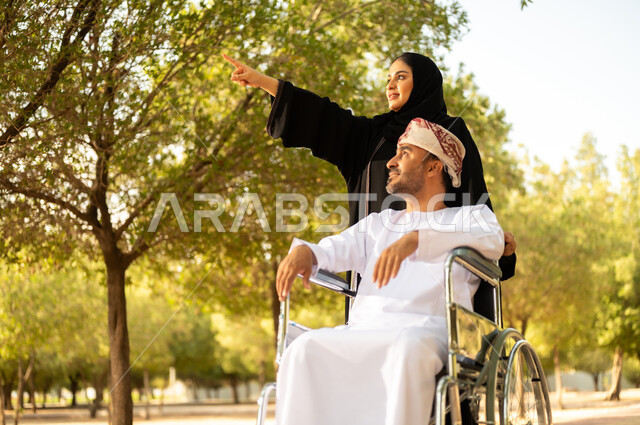 مساعدة مرضى الإعاقة الحركية على التنقل بسهولة، الاستمتاع وقضاء وقت ممتع بالهواء الطلق، العطف والحب بين أفراد الأسرة, امرأة عربية خليجية عمانية محجبة ترتدي العباءة تتنزه في الحديقة برفقة زوجها المقعد, شعور ذوي الهمم بالراحة والطمأنينة في بيئتهم